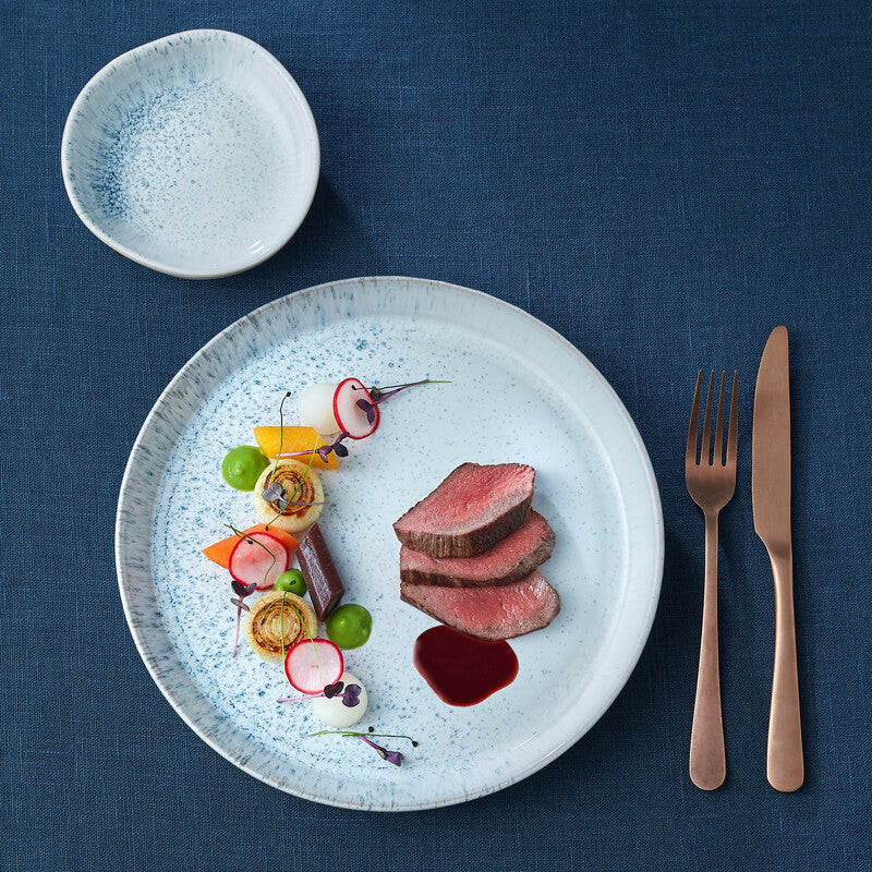 Lifestyle image of Kiln Blue dinner plate and small coupe bowls on a Blue tablecloth